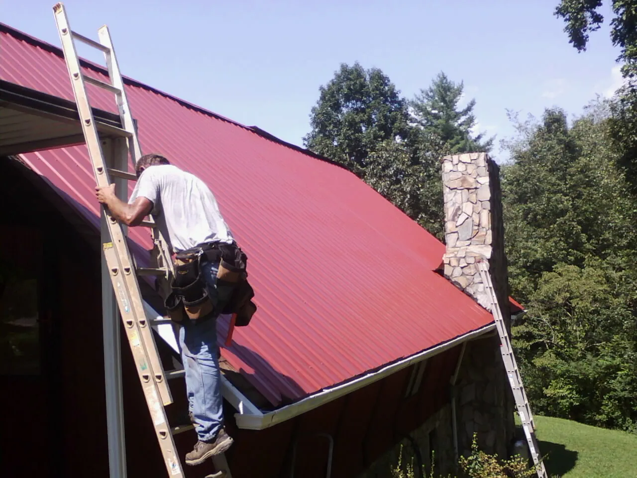 Roofer on a ladder beside a red metal roof and stone chimney — Soffit & Fascia Repair by Roofing Brothers in Pevely