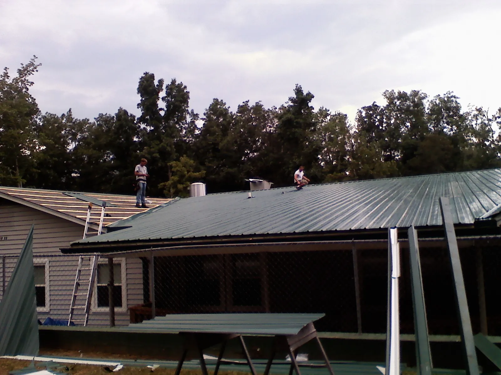 Two roofers installing green metal panels above a covered porch in Pevely